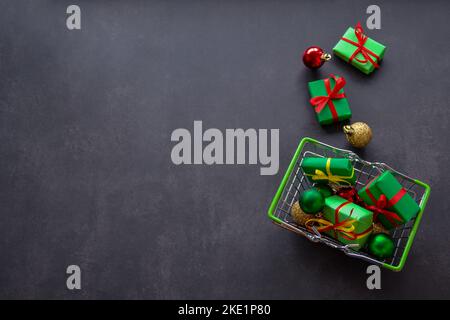 Warenkorb mit Geschenken in grünem Papier, roten und gelben Schleifen, Weihnachtskugeln aus rot, grün, Gold auf schwarzem Hintergrund. Einige Geschenke, Kugeln liegen in der Nähe. Stockfoto