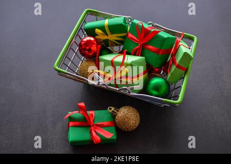 Warenkorb mit Geschenken in grünem Papier, roten und gelben Schleifen, Weihnachtskugeln aus rot, grün, Gold auf schwarzem Hintergrund. Einige Geschenke, Kugeln liegen in der Nähe. Stockfoto