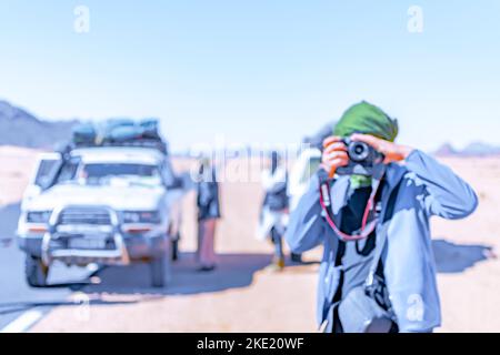 Ein verschwommener Mann, der in der Wüste Fotos mit einer DSLR macht, mit einem verschwommenen Hintergrund eines Allradwagens und Tuareg-Menschen. Stockfoto