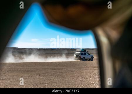 Fensterblick auf ein Geländefahrzeug im Geländewagen, das in einem Sandsteingelände mit Staub dahinter, felsigen Wüstenbergen und blauem Himmel rasiert. Stockfoto