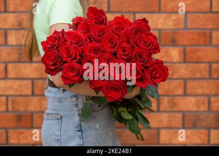 Frau mit einem luxuriösen Strauß frischer roter Rosen vor dem Hintergrund einer Ziegelmauer. Mono Bouquet von roten Rosen in den Händen der Frau. Stockfoto