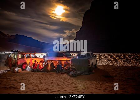 Campinggruppe in der Sahara-Wüste bei Nacht. GELÄNDEWAGEN geparkt, Touristen und lokale Berber am Lagerfeuer sitzen. wolkiger Himmel Mondschein. Stockfoto