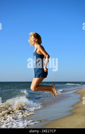 Junge, schöne Frau aus weißer Rennblonde springt vor dem Hintergrund der Wellen am Strand und lacht fröhlich Stockfoto