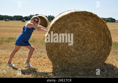 Eine junge Frau in Sonnenbrillen lächelt scherzhaft schiebt einen runden Heuhaufen in ein großes Getreidefeld Stockfoto