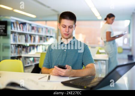Ein junger Student, der an einem Schreibtisch in der Universitätsbibliothek sitzt, schreibt mit Freunden und fotografiert auf dem Handy Stockfoto