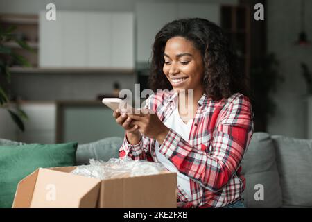 Glückliche hübsche schwarze junge Frau Client in lässig auspackt Paket, macht Foto von Käufen auf dem Smartphone Stockfoto