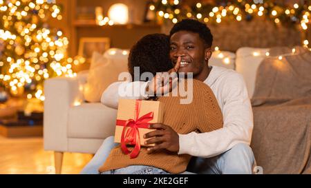 Glückliche junge schwarze Frau umarmen Mann mit Geschenkbox, Mann zeigen shh Zeichen in gemütlichen Wohnzimmer Interieur Stockfoto