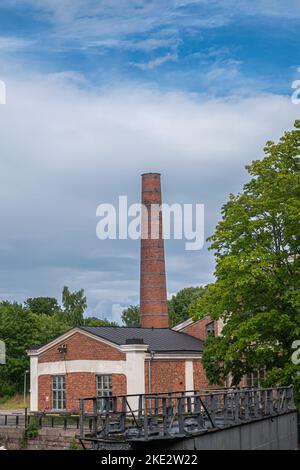 Helsinki, Finnland - 19. Juli 2022: Festung Suomenlinna. Lavyhalli-Gebäude mit hohem roten Ziegelkamin vor blauer Wolkenlandschaft. Grünes Laub und Wat Stockfoto