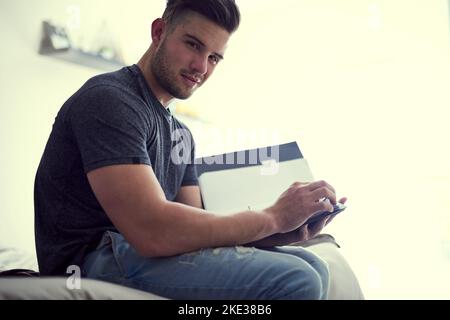Jeder Erfolg beginnt mit der Entscheidung, es zu versuchen. Porträt eines selbstbewussten jungen Mannes, der zu Hause auf seinem Bett sitzt und Papierkram studiert. Stockfoto