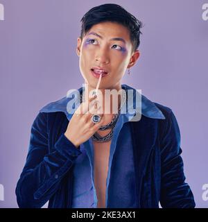 Beauty, Make-up und lgbt Asian Mann im Studio auf lila Hintergrund mit Kosmetik, Beauty-Produkte und Lippenstift. Kreativ, Mode und Queer oder lgbtq Stockfoto