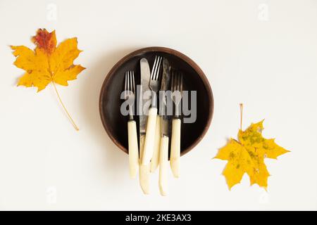 Messer und Gabeln liegen in einer tiefen Tonplatte, die im Herbst bei trockenen Ahornblättern aus nächster Nähe auf weißem Hintergrund steht Stockfoto