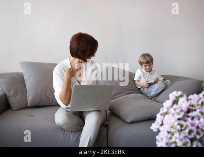 Mutter und Sohn mit digitalen Gadgets sitzen auf dem Sofa im Wohnzimmer. Unkenntliche Frau arbeitet zu Hause auf einem Laptop, neben einem Kind spielt Stockfoto
