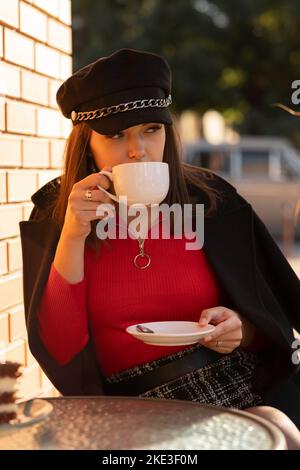 Nachdenkliches Mädchen in stilvollem schwarzen Hut und herbstlich leuchtendem roten Pullover, das in einem Pariser Café im Freien sitzt und Kaffee trinkt. Stockfoto