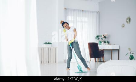 Glückliche Hausfrau macht Hausarbeit und hört Musik mit modernen kabellosen Kopfhörern, hübsche junge Dame singt im Mopp und tanzt mit Spaß zu Hause. Stockfoto