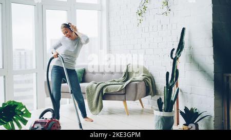 Die fröhliche blonde Hausfrau verwendet den Staubsauger während der routinemäßigen Säuberung zu Hause, hört Musik über Kopfhörer, singt und tanzt. Technologie und junge Menschen Konzept. Stockfoto