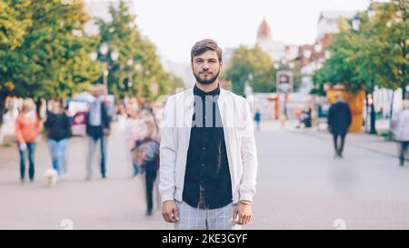 Porträt eines ernsthaften jungen Mannes, der im Sommer in stilvoller Kleidung im Zentrum der belebten Fußgängerzone die Kamera ansieht, während viele Menschen vorbeikommen. Stockfoto
