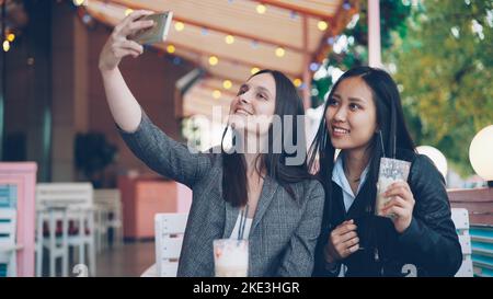 Fröhliche Freundinnen führen Online-Videoanruf mit einem modernen Smartphone, das auf den Bildschirm schaut und im Straßencafé miteinander redet. Kommunikations- und Technologiekonzept. Stockfoto