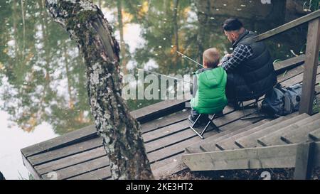 Aus der Perspektive von Menschen, die Vater und Sohn im Teich im Wald fischen, sitzen auf Stühlen am hölzernen Pier und halten Ruten. Familie, gemeinsames Hobby und Naturkonzept. Stockfoto