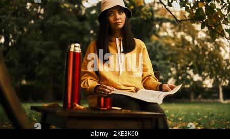 Ein Mädchen sitzt auf einem Stuhl im Park, trinkt Tee und liest ein Buch Stockfoto