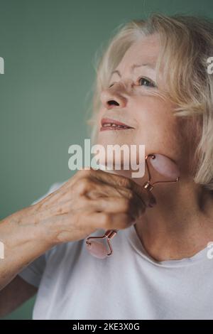 Nahaufnahme einer älteren Frau, die mit einem Jaderollen eine Gesichtsmassage macht und dabei die Augen schließt. Hübsche Frau putzen frische Haut. Antiage-Behandlung Stockfoto