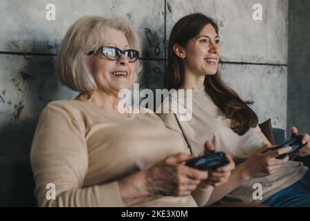 Aufgeregt konzentrierte ältere Mutter mit ihrer erwachsenen Tochter, die Videokonsolenspiele spielt, während sie im Wohnzimmer zu Hause auf dem Sofa sitzt, lacht und Stockfoto