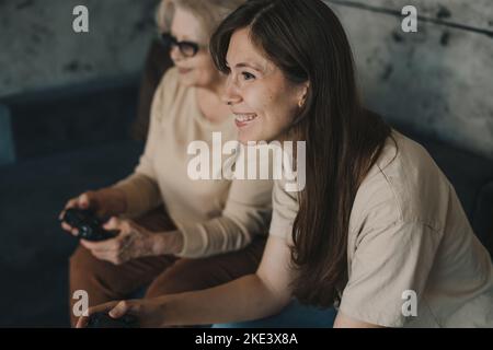 Ältere Großmutter spielt mit ihrer Tochter Videospiele mit Konsole, jubelende Sieg, Hobby. Lachen und Spaß haben. Freizeit und Menschen Stockfoto
