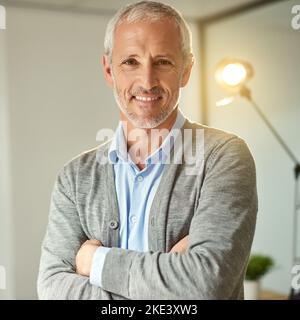 Erfolg im Schritt. Porträt eines lächelnden, reifen Geschäftsmannes, der mit gekreuzten Armen in einem Büro steht. Stockfoto