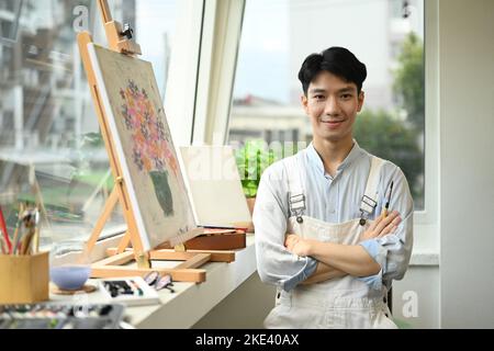 Porträt eines hübschen männlichen Künstlers, der mit gekreuzten Armen vor der Staffelei im hellen Kunststudio steht und vor der Kamera lächelt Stockfoto