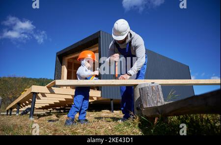 Vater mit Kleinkind Sohn Gebäude Holzrahmen Haus. Männliche Bauherren, die auf der Baustelle Nagel in die Planke schlagen, tragen an sonnigen Tagen Helm und blaue Overalls. Zimmerei- und Familienkonzept. Stockfoto