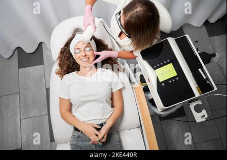 Moderne Methode Laser-Entfernung von problematischen Teleangiektasen auf dem Gesicht. Draufsicht Zufriedene lockige Mädchen auf Verfahren der Behandlung vaskulären Sternchen Gesicht per Laser. Stockfoto