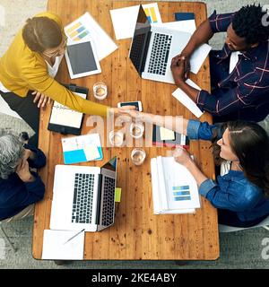 Willkommen an unserem Tisch. Ein Team von Kreativen, die sich gegenseitig die Hände über einen Tisch schüttelten, hat eine Aufnahme in einem hohen Winkel gemacht. Stockfoto