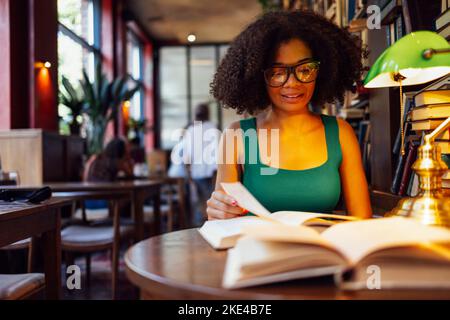 Glückliche afroamerikanische Studentin, die in der Universitätsbibliothek studding und auf das Buch schaute, in einer Brille mit grüner Lampe. Lächelnd teen Mädchen in gemütlich Stockfoto