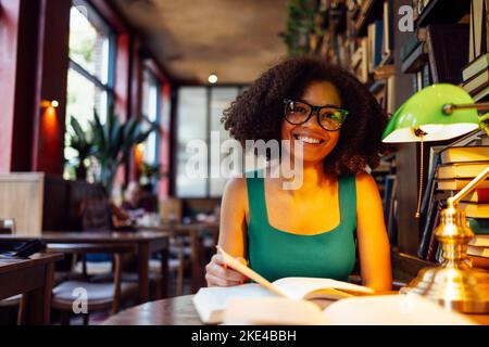 Glückliche afroamerikanische Studentin, die in der Universitätsbibliothek studding und die Kamera ansah und eine Brille an der grünen Lampe trug. Lächelndes Teenager-Mädchen im gemütlichen li Stockfoto