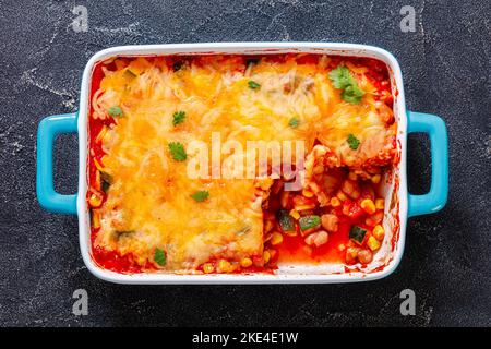 Vegetarische Enchilada-Kasserolle mit Tortillas, Sauce, Pinto-Bohnen, Mais, Zucchini und mexikanischem Käse in einer Auflaufform auf einem Betontisch, Stockfoto