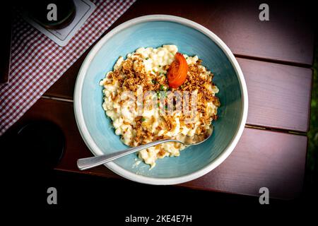 Eine deutsche Spezialität - Allgaeuer Kaesespätzle mit Zwiebeln und Tomaten in einer Schüssel auf einem Holztisch. Stockfoto