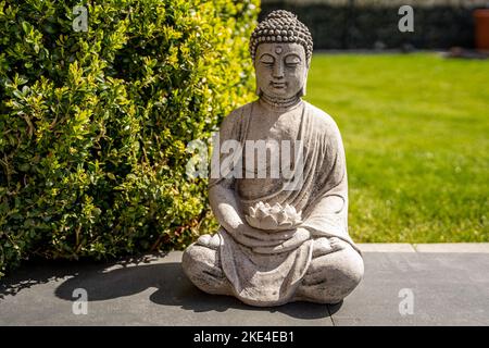 Buddha-Statue, die neben einem Busch vor dem Gras sitzt Stockfoto