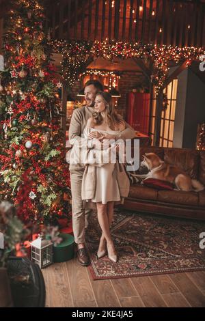Porträt eines jungen schönen Paares umarmt Indoor Eve 25 Dezember mit akita inu Hund. Menschen in Strick stilvolle Outfit Umarmungen küsst Zärtlichkeit feiert ne Stockfoto