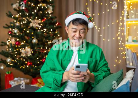 Ein junger asiatischer Mann in einem roten Weihnachtsmannhut und einem grünen Hemd hält ein Telefon in den Händen, schreibt eine Nachricht und gratuliert zu den Weihnachtsferien. Sitzen zu Hause auf dem Sofa in der Nähe des Weihnachtsbaums. Stockfoto