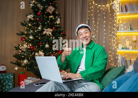 Porträt eines jungen, hübschen asiatischen Mannes, der zu Hause in der Nähe des Weihnachtsbaums in einem roten Hut auf dem Sofa sitzt, einen Laptop und ein Telefon in der Hand hält, lächelnd auf die Kamera blickt und super zeigt. Stockfoto
