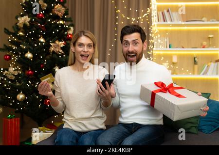 Online-Shopping. Glückliches junges Paar zu Hause in der Nähe von Weihnachtsbaum, mit Kreditkarte, Telefon und Geschenk. Wir bekamen ein unerwartetes Weihnachtspäckchen. Stockfoto