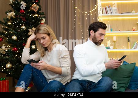 Ein junges Ehepaar, ein Mann und eine Frau, an den Weihnachts- und Neujahrsferien. Sie sitzen zu Hause auf dem Sofa, die Beleidigten haben sich den Rücken gekehrt, schauen auf ihre Telefone Stockfoto