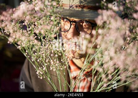 Porträt eines attraktiven kaukasischen Mannes mit Brille und Hut, der in einem frischen Blumenstrauß taucht. Konzept zum Muttertag, Valentinstag oder Internationalen Frauentag. Hochwertige Fotos Stockfoto