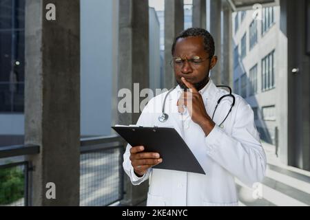Ernst und denken reifen afroamerikanischen Arzt außerhalb der modernen Klinik, Denken und Lesen Behandlung Bericht und Dokumente. Stockfoto