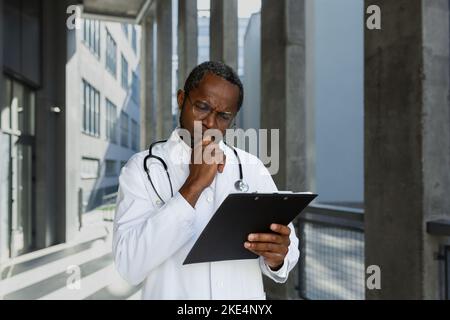 Ernst und denken reifen afroamerikanischen Arzt außerhalb der modernen Klinik, Denken und Lesen Behandlung Bericht und Dokumente. Stockfoto