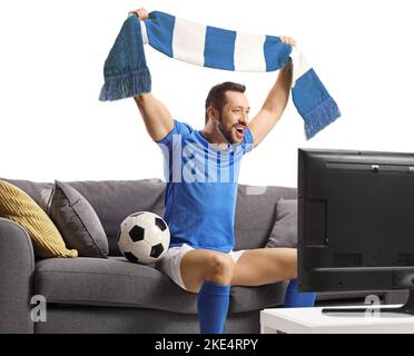 Fröhlicher Mann in einem Fußballtrikot, der mit einem Schal jubelt und auf einem Sofa vor einem auf weißem Hintergrund isolierten fernseher sitzt Stockfoto