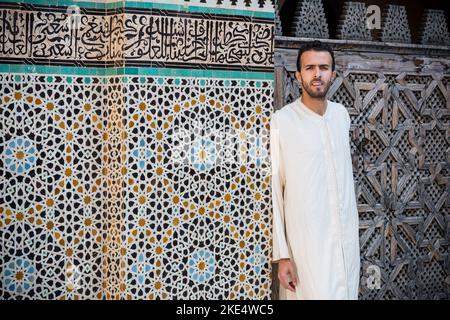 Junge muslimische Mann in traditioneller Kleidung tragen lange weiße Hemd vor der Wand mit Text aus Koran und arabesque Dekoration mit Flo Stockfoto