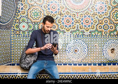 Junger muslimischer Mann, der ein Mobiltelefon benutzt, das vor einem traditionellen arabesken Mosaik sitzt Stockfoto