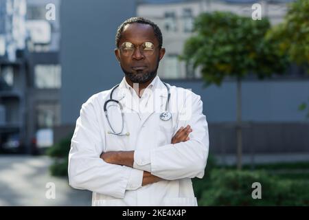 Porträt eines ernsthaft denkenden afroamerikanischen Arztes, eines Mannes in einer Brille und eines medizinischen Mantels mit Stethoskop, der außerhalb der modernen Klinik mit gekreuzten Armen auf die Kamera blickt. Stockfoto