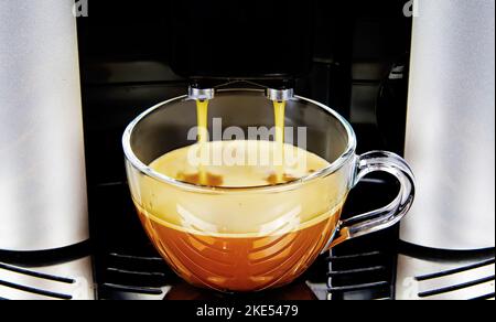 Nahaufnahme des Ausgangs einer modernen vollautomatischen Kaffeemaschine, eine isolierte, transparente Glasbecherfüllung mit frisch gebrühter Café-Crema Stockfoto