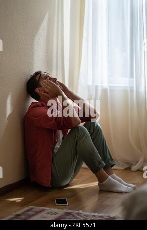 Aufgeregter, depressiver junger Mann, der zu Hause auf dem Boden sitzt, erhielt unerwartete schlechte Nachrichten über das Telefon Stockfoto